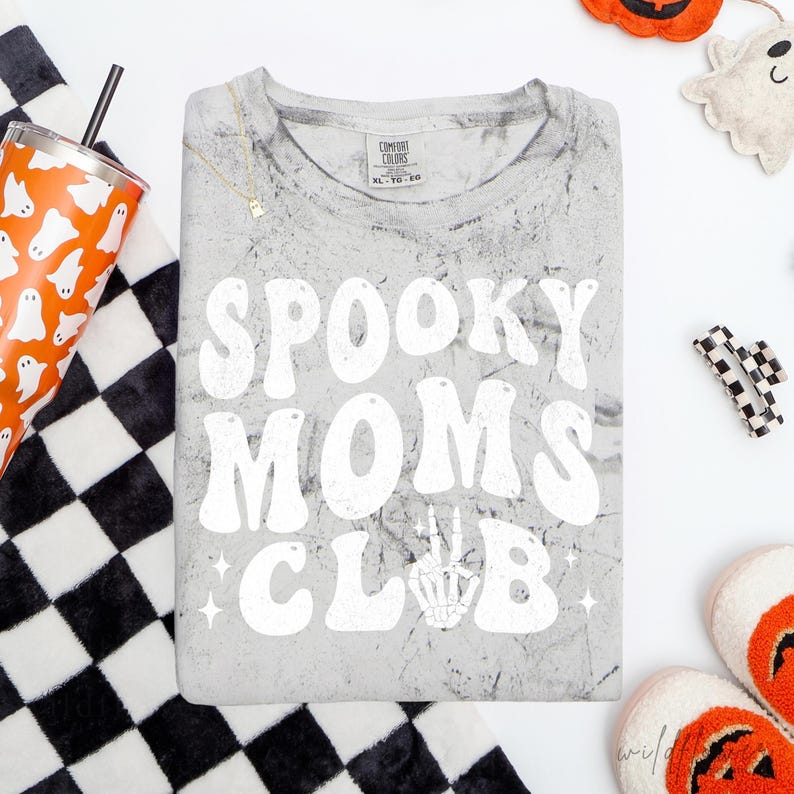 May include: A grey acid-wash t-shirt featuring the white text SPOOKY MOMS CLUB in a retro font, with a skeleton hand making a peace sign replacing the letter M. The shirt has a crew neckline and a Comfort Colors brand label at the inner collar.