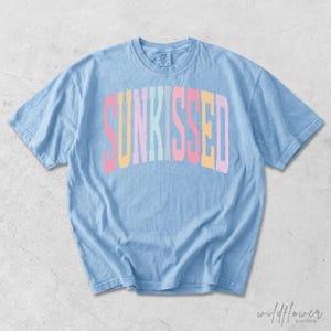 May include: A light blue short-sleeve t-shirt featuring the word SUNKISSED printed in an arched, multi-colored font with letters in shades of pink, yellow, and light blue. The garment has a crew neckline and a relaxed, casual fit.