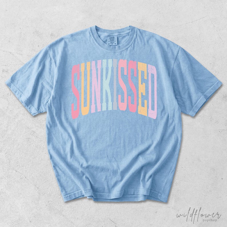 May include: A light blue short-sleeve t-shirt featuring the word SUNKISSED printed in an arched, multi-colored font with letters in shades of pink, yellow, and light blue. The garment has a crew neckline and a relaxed, casual fit.