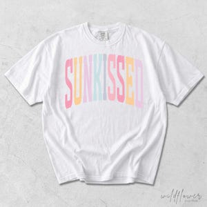 May include: A white short-sleeve t-shirt featuring the word SUNKISSED printed in a large, arched, multi-colored font across the chest. The letters alternate between shades of pink, yellow, light blue, and lavender in a block-style typeface.
