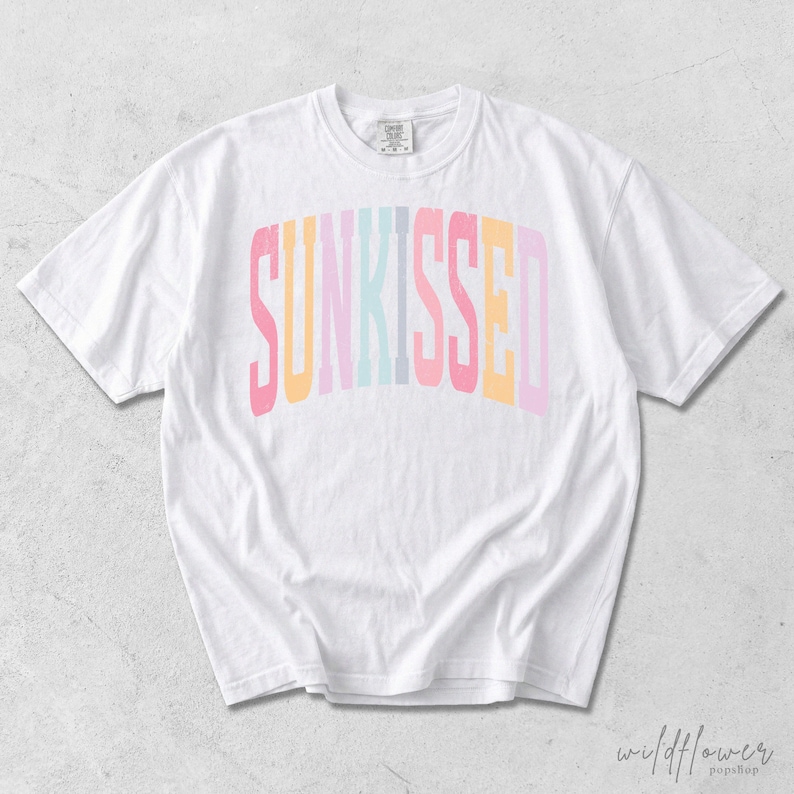 May include: A white short-sleeve t-shirt featuring the word SUNKISSED printed in a large, arched, multi-colored font across the chest. The letters alternate between shades of pink, yellow, light blue, and lavender in a block-style typeface.