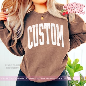 May include: A brown long-sleeve crewneck sweatshirt featuring the word CUSTOM printed in large, white, distressed-style capital letters across the chest. The garment has a relaxed fit with ribbed cuffs and a ribbed neckline.