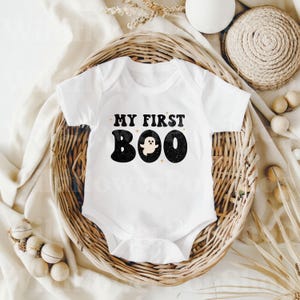 May include: A white short-sleeved baby bodysuit featuring the black text MY FIRST BOO with a small ghost graphic replacing the letter O. The design has a distressed texture and is accented with small four-pointed stars around the lettering.