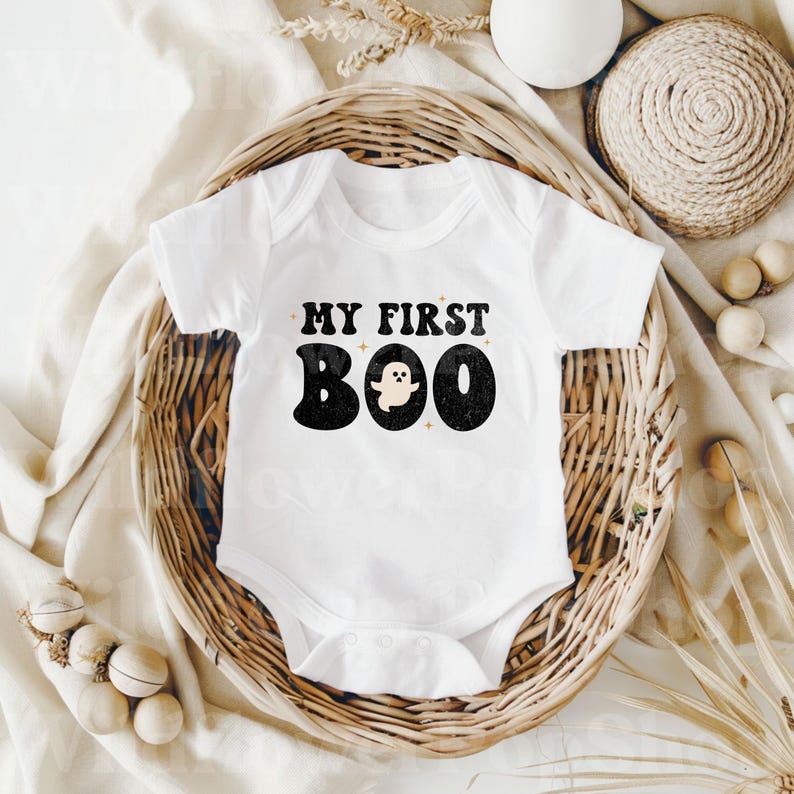 May include: A white short-sleeved baby bodysuit featuring the black text MY FIRST BOO with a small ghost graphic replacing the letter O. The design has a distressed texture and is accented with small four-pointed stars around the lettering.