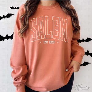May include: A terracotta orange long-sleeve crewneck sweatshirt featuring the white text SALEM in a collegiate font, with the text EST 1626 centered below it, flanked by two small white stars on either side of the date.