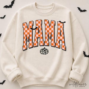 Puede incluir: Una sudadera de cuello redondo color crema con la palabra MAMA en una fuente universitaria grande a cuadros naranja y rosa claro. El dise&ntilde;o incluye peque&ntilde;as siluetas de murci&eacute;lagos negros y un gr&aacute;fico de calabaza con estampado de vaca.