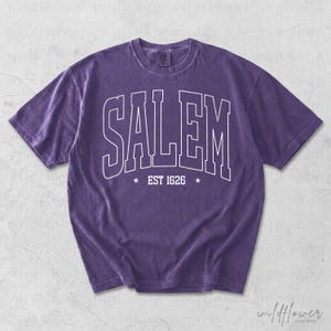 May include: A purple short-sleeve t-shirt featuring the white outlined text SALEM in an arched collegiate font across the chest, with the text EST 1626 centered below between two small white stars. The fabric has a washed, vintage appearance.
