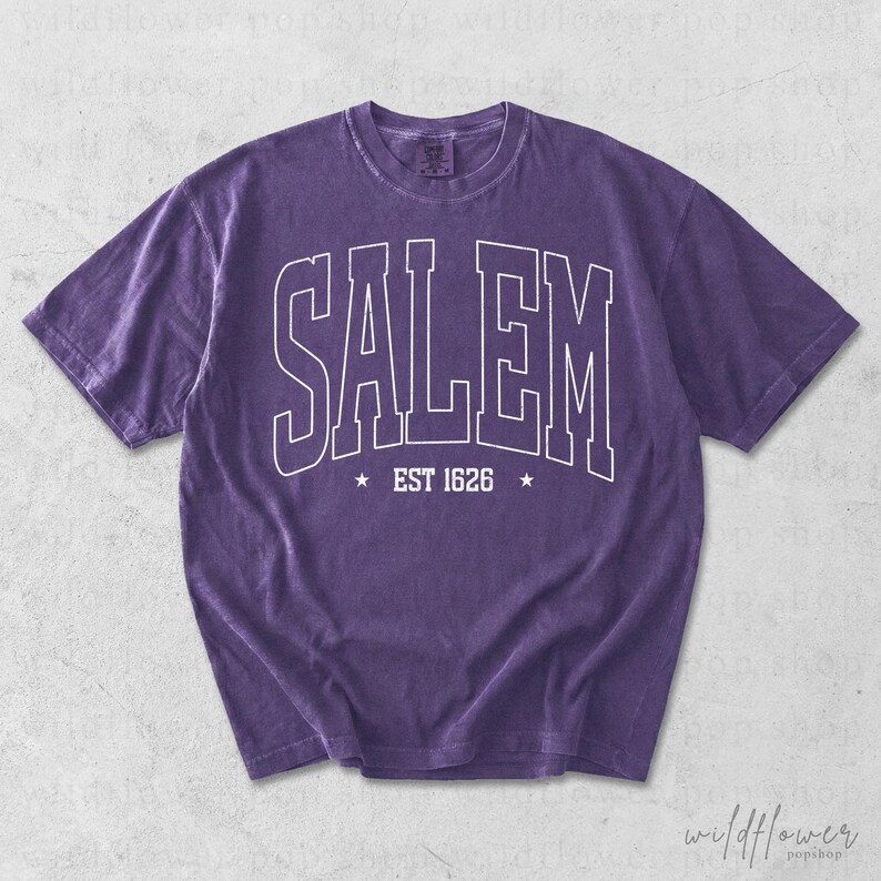 May include: A purple short-sleeve t-shirt featuring the white outlined text SALEM in an arched collegiate font across the chest, with the text EST 1626 centered below between two small white stars. The fabric has a washed, vintage appearance.