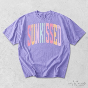 May include: A light purple short-sleeve t-shirt featuring the word SUNKISSED printed in a large, arched, multi-colored font across the chest. The letters are colored in shades of pink, yellow, light blue, and white, with a distressed texture.