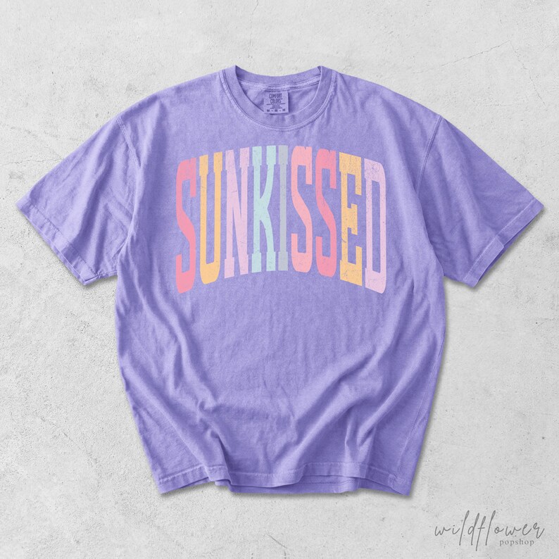 May include: A light purple short-sleeve t-shirt featuring the word SUNKISSED printed in a large, arched, multi-colored font across the chest. The letters are colored in shades of pink, yellow, light blue, and white, with a distressed texture.