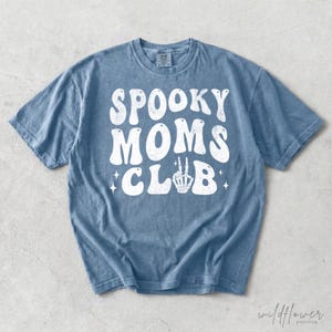 May include: A blue short-sleeve t-shirt with a distressed white graphic print on the front. The text reads SPOOKY MOMS CLUB in a bold, wavy font, with a skeleton hand making a peace sign replacing the letter L and small star accents.