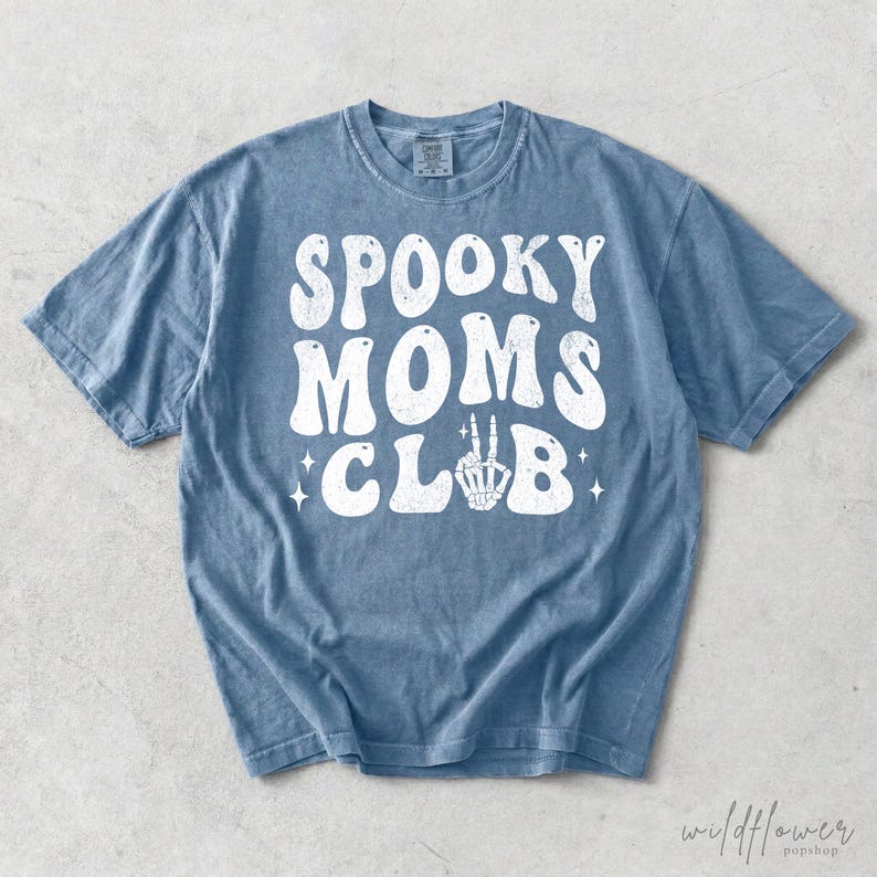 May include: A blue short-sleeve t-shirt with a distressed white graphic print on the front. The text reads SPOOKY MOMS CLUB in a bold, wavy font, with a skeleton hand making a peace sign replacing the letter L and small star accents.