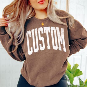 Könnte beinhalten: Ein braunes Sweatshirt mit dem Wort "CUSTOM" in großen, weißen Blockbuchstaben auf der Brust. Das Sweatshirt hat lange Ärmel und einen Rundhalsausschnitt. Der Stoff scheint ein weiches, bequemes Material zu sein.