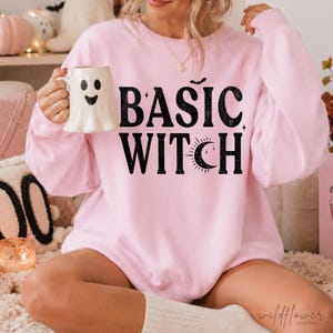 Può includere: Una felpa girocollo rosa chiaro con la scritta nera BASIC WITCH, una piccola icona di pipistrello sopra il testo e un'icona di mezzaluna integrata nella lettera T. La felpa è indossata con una tazza in ceramica bianca a forma di fantasma.