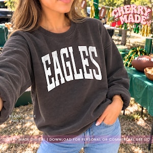 Peut inclure: Un sweat-shirt à col rond gris anthracite arborant le mot EAGLES imprimé en grandes lettres blanches à l'aspect vieilli sur la poitrine. Le vêtement présente des manches longues ainsi qu'un col, des poignets et un ourlet côtelés.