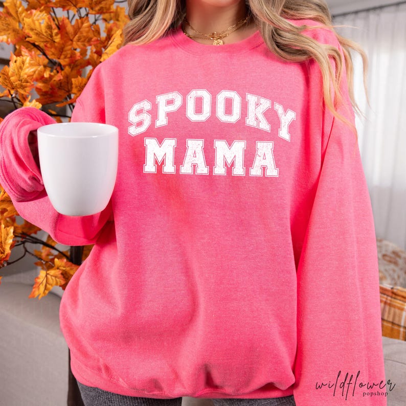 May include: A bright pink long-sleeve sweatshirt featuring the white text SPOOKY MAMA in a collegiate font across the chest. The garment has a crew neckline and long sleeves, and it is paired with a plain white ceramic mug held in one hand.