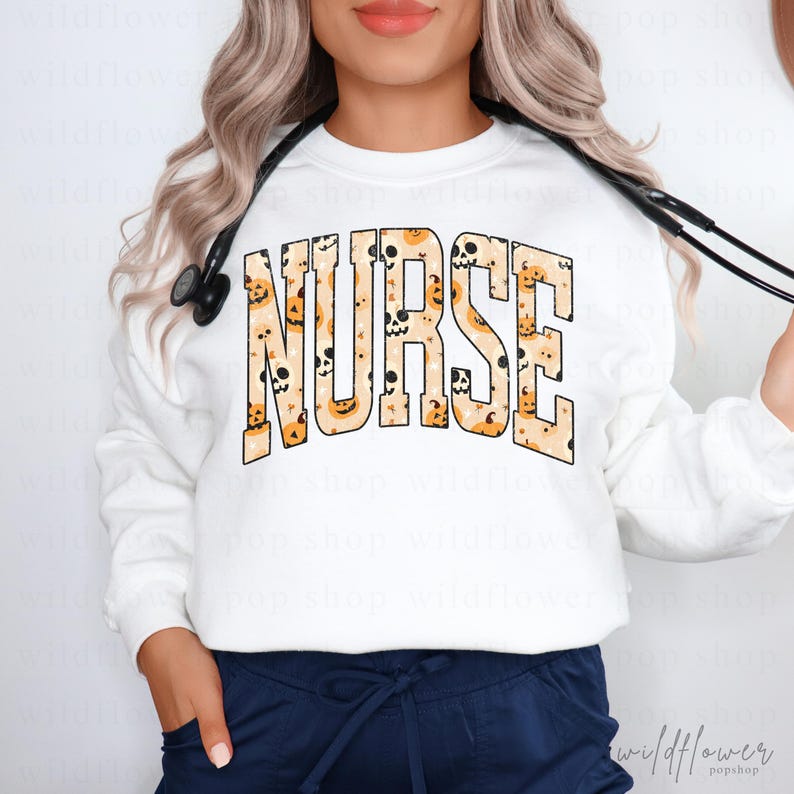 May include: A white long-sleeve sweatshirt featuring the word NURSE in large, block-style letters. The letters are filled with a repeating pattern of orange pumpkins and white skulls. A black stethoscope is draped around the neck of the garment.