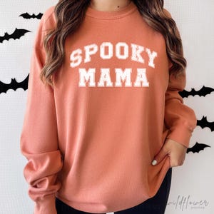 May include: A long-sleeve crewneck sweatshirt in a muted orange color. The front features the text SPOOKY MAMA printed in a distressed white collegiate-style font. The garment has a ribbed neckline, cuffs, and hem, providing a classic fit.
