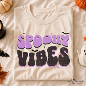 May include: A cream-coloured t-shirt featuring the text SPOOKY VIBES in a retro, wavy font. The word SPOOKY is in light purple, and VIBES is in black with a distressed texture. Small black bats and purple star accents surround the lettering.