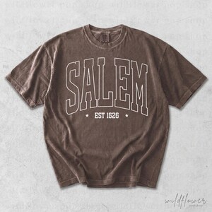 May include: A brown short-sleeve t-shirt featuring the white text SALEM in a large arched collegiate font across the chest, with the text EST 1626 centered below it between two small white stars. The shirt has a crew neckline and short sleeves.