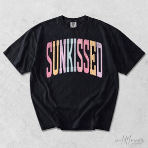 May include: A black short-sleeve t-shirt featuring the word SUNKISSED printed in a large, arched, distressed font across the chest. The letters alternate between pastel pink, orange, light blue, and yellow colors, creating a vibrant contrast.