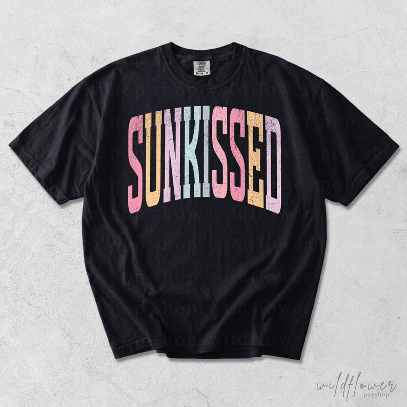 May include: A black short-sleeve t-shirt featuring the word SUNKISSED printed in a large, arched, distressed font across the chest. The letters alternate between pastel pink, orange, light blue, and yellow colors, creating a vibrant contrast.