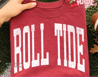 May include: A maroon crew neck t-shirt featuring the white distressed text ROLL TIDE printed in a large arched font across the chest. The shirt includes a small logo in the top right corner with the text CHERRY MADE DIGITAL and two cherries.