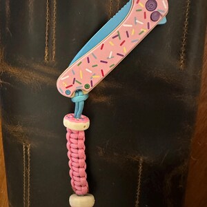 Op de afbeelding: Een roze en blauw zakmes met een donut-thema. Het mes heeft een roze handvat met witte en gekleurde sprinkles. Het mes is bevestigd aan een roze paracord-koord met witte donutvormige kralen.