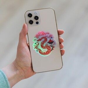 Pu&ograve; includere: Un iPhone bianco con un adesivo olografico di un serpente rosso con un pagoda e fiori di ciliegio. L'adesivo ha caratteri cinesi.