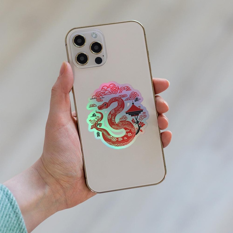 Pu&ograve; includere: Un iPhone bianco con un adesivo olografico di un serpente rosso con un pagoda e fiori di ciliegio. L'adesivo ha caratteri cinesi.