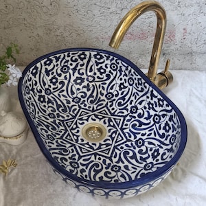 Op de afbeelding: Een ovale keramische waskom met een blauw-wit bloemmotief, een centraal stermotief en een goudkleurige metalen kraan. De waskom heeft een gladde, afgeronde rand en een metalen afvoer in het midden van het gedetailleerde blauwe ontwerp.