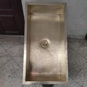 Op de afbeelding: Een rechthoekige, gehamerde koperen spoelbak met een centrale afvoer. De spoelbak heeft een gepolijste, gouden afwerking en een getextureerd oppervlak. Het ontwerp is eenvoudig en modern, geschikt voor een keuken of badkamer.