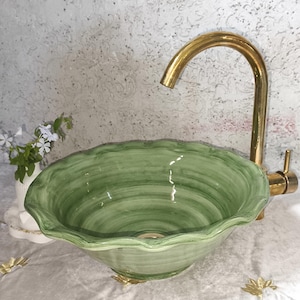 Op de afbeelding: Een ronde keramische waskom met een saliegroene afwerking, zichtbare penseelstreektexturen en een geschulpte, golvende rand. De waskom is gecombineerd met een hoge, gebogen, goudkleurige metalen kraan met een enkele hendel.