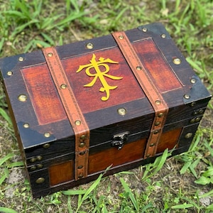 May include: A wooden chest with a dark brown exterior and a lighter brown top. The chest features leather straps, metal corner protectors, and a gold emblem on the lid. The chest is sitting on green grass.
