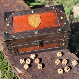 May include: A wooden treasure chest with brass accents and a leather emblem, accompanied by several dice. The chest has a reddish-brown top and a darker brown frame. The dice are light-colored with black dots.