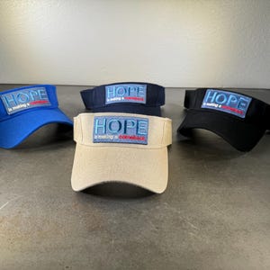 May include: Four visors with the word "HOPE" in blue lettering and the phrase "is making a comeback" in red lettering on a patch on the front of each visor. The visors are in blue, black, beige, and navy blue.