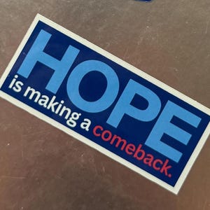 Peut inclure: Un autocollant blanc avec le mot "HOPE" en lettres bleues et rouges. Le texte "is making a comeback." est écrit en lettres rouges plus petites sous le mot "HOPE".