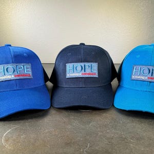 May include: Three baseball caps in blue, navy blue, and turquoise. Each cap has a patch with the word "HOPE" and the text "is making a comeback" in red and white.