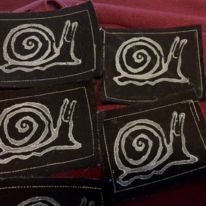 Könnte beinhalten: Eine Sammlung rechteckiger schwarzer Stoffaufnäher, die jeweils mit einer weißen Siebdruckgrafik einer Schnecke mit Spiralgehäuse versehen sind. Die Aufnäher haben sichtbare weiße Nähte an den Rändern, die einen Rahmen um das Schneckenmotiv bilden.