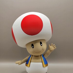 May include: A figurine of the character Toad from the Super Mario video game series. The figure has a large white mushroom cap with a red circle on top. He wears a blue vest with gold trim, a white vest, and brown shoes. Toad is waving.