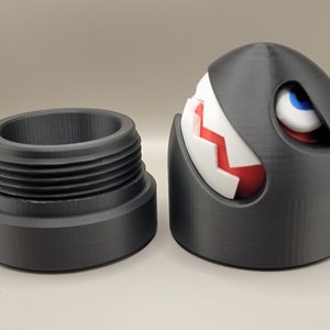 May include: A 3D-printed, black, cartoon-style character with a white and red segmented design, blue eye, and a separate threaded base. The character is a stylized, spherical design with a menacing expression. The base is cylindrical.