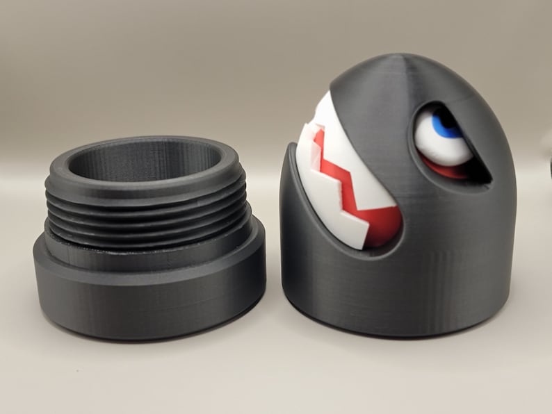 May include: A 3D-printed, black, cartoon-style character with a white and red segmented design, blue eye, and a separate threaded base. The character is a stylized, spherical design with a menacing expression. The base is cylindrical.