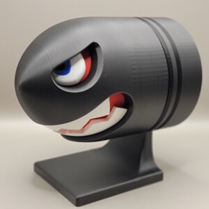 May include: A black Bullet Bill figure with a menacing expression, featuring a white-toothed grin, red mouth, and a single eye with blue, white, and red details. The figure is mounted on a black base.