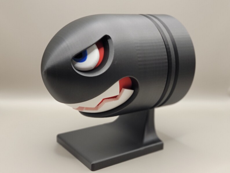May include: A black Bullet Bill figure with a menacing expression, featuring a white-toothed grin, red mouth, and a single eye with blue, white, and red details. The figure is mounted on a black base.