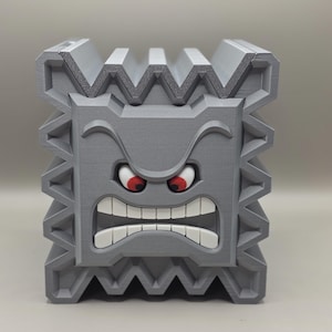 May include: A gray, 3D-printed Thwomp from the Super Mario video game series. The block-shaped character has an angry expression with red eyes and a wide, toothy grin. The edges of the block are jagged.