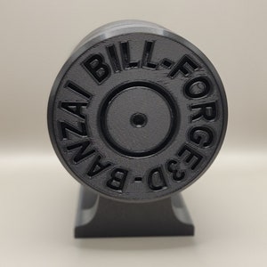 May include: A black 3D-printed object with the text "BILL-FORGED 3D-BANZAI" in a circular pattern. The object is cylindrical and mounted on a black base, showcasing a textured surface.