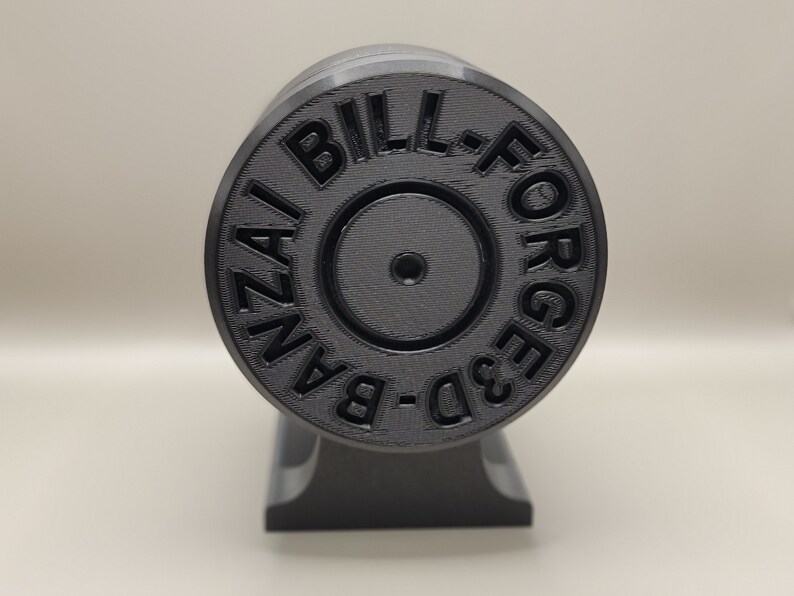 May include: A black 3D-printed object with the text "BILL-FORGED 3D-BANZAI" in a circular pattern. The object is cylindrical and mounted on a black base, showcasing a textured surface.