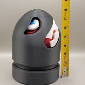May include: A gray Bullet Bill figure, approximately 7 inches tall, with a white and red mouth and a blue-eyed gaze. The figure has a cylindrical base and a rounded top, resembling a cartoon character.