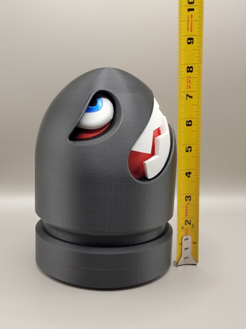 May include: A gray Bullet Bill figure, approximately 7 inches tall, with a white and red mouth and a blue-eyed gaze. The figure has a cylindrical base and a rounded top, resembling a cartoon character.