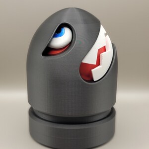 May include: A gray, 3D-printed Bullet Bill figure, a character from the Super Mario video game series. The figure has a white and red mouth, a blue and white eye, and a cylindrical base.